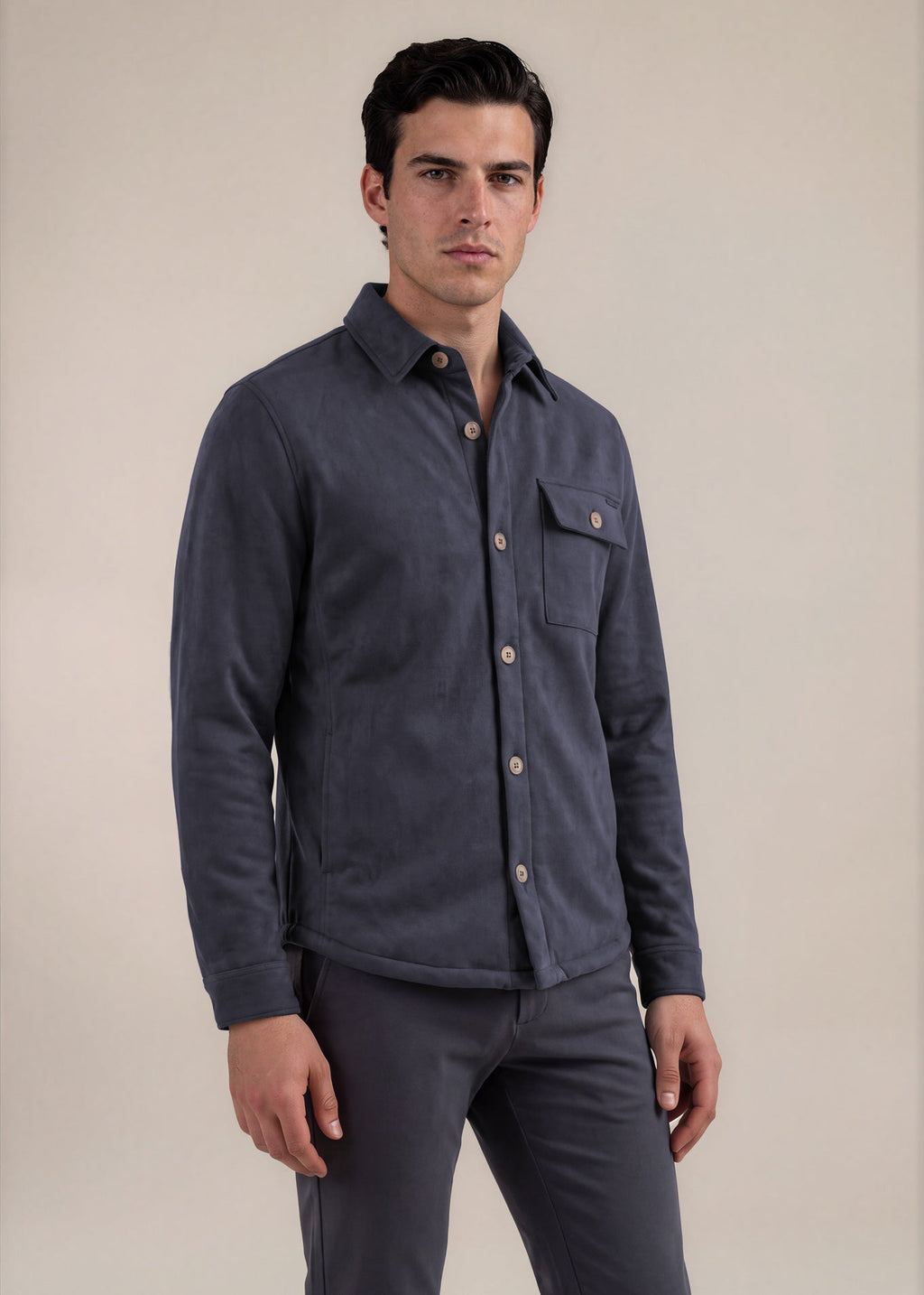 A man wearing a dark slate blue button-up shirt jacket with a chest pocket and matching dark trousers, standing against a plain beige studio background.