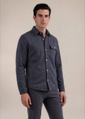 A man wearing a dark slate blue button-up shirt jacket with a chest pocket and matching dark trousers, standing against a plain beige studio background.
