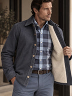 A man wearing a navy overshirt shirt jacket with a cream sherpa lining over a blue and white plaid button-down shirt, paired with gray trousers and a brown belt.