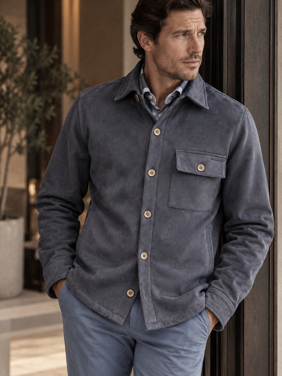 A man wearing a dark navy sueded button-up shirt jacket over a light blue collared shirt, paired with blue trousers, standing in a softly lit indoor setting.