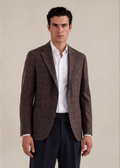 A man wearing a brown and gray checkered blazer over a white dress shirt and navy trousers, standing against a plain beige studio background.