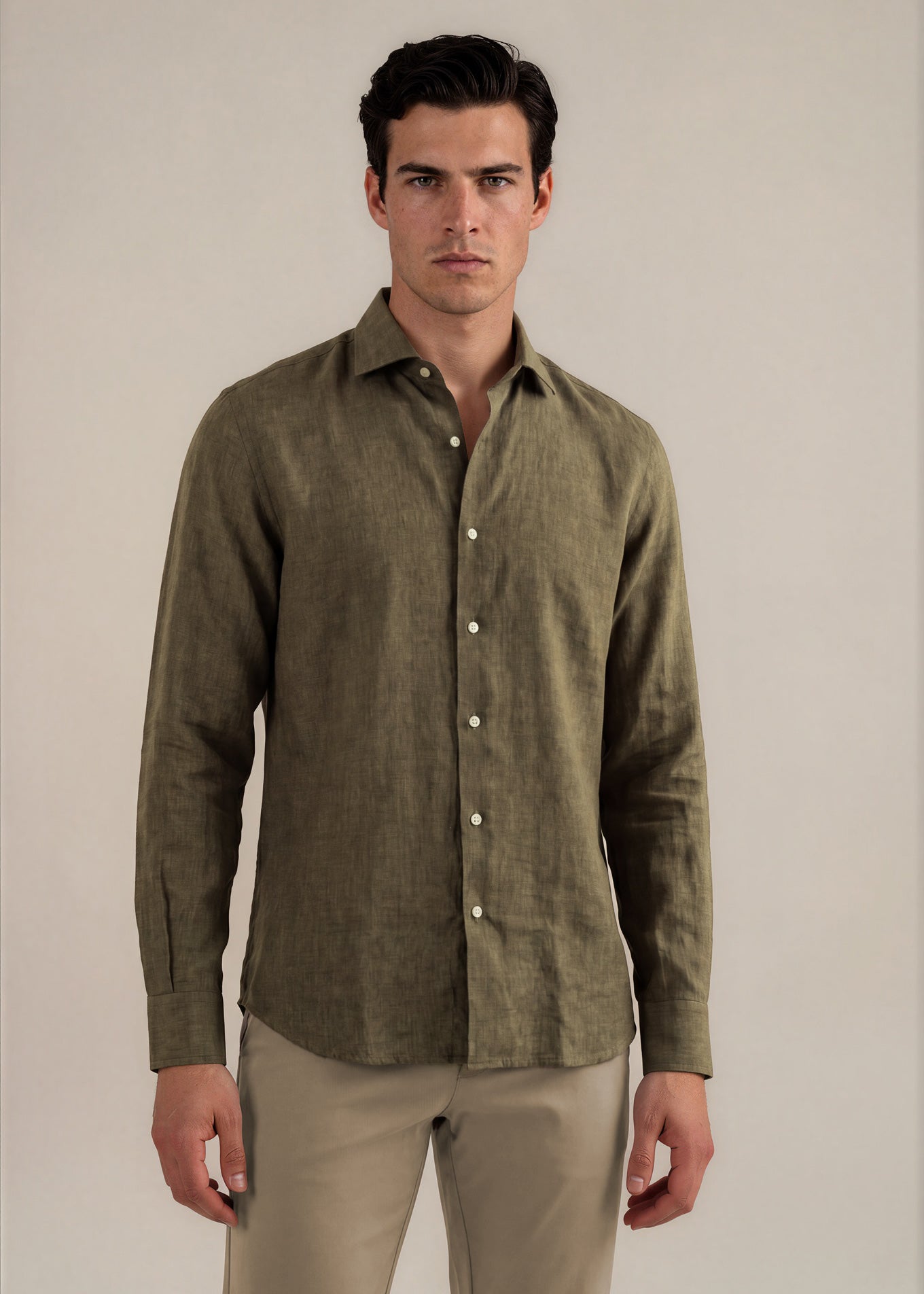 A man wearing a textured olive green linen button-down shirt and beige trousers, standing against a plain light beige studio background.