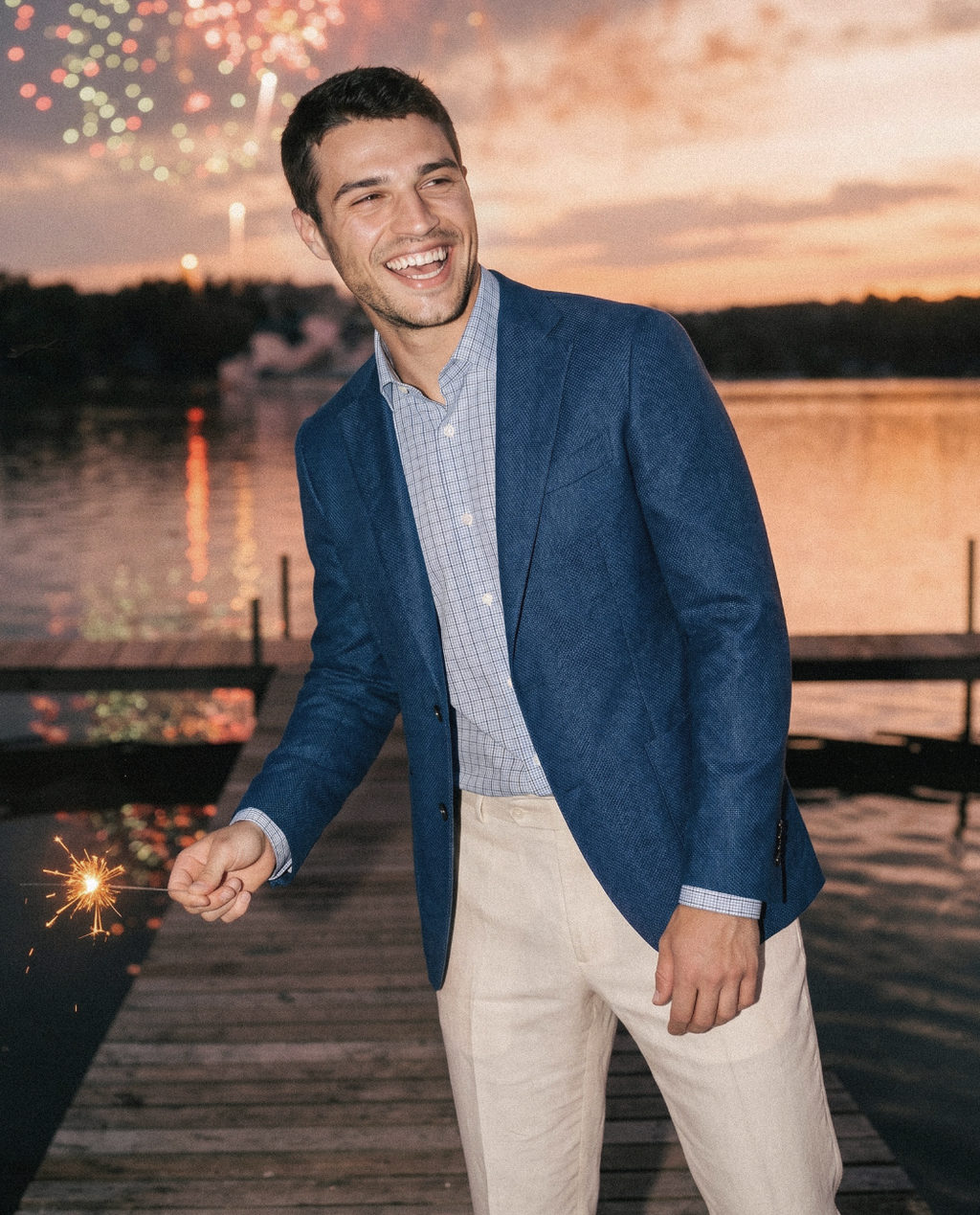 Man in a blue blazer holding a sparkler by a lake with fireworks in the background