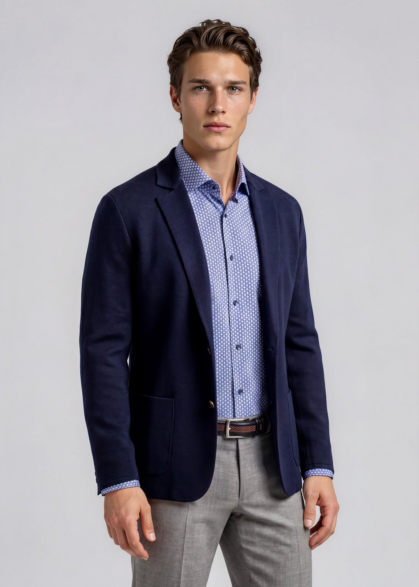 Man wearing a navy blazer, light blue patterned shirt, and gray pants on a plain background