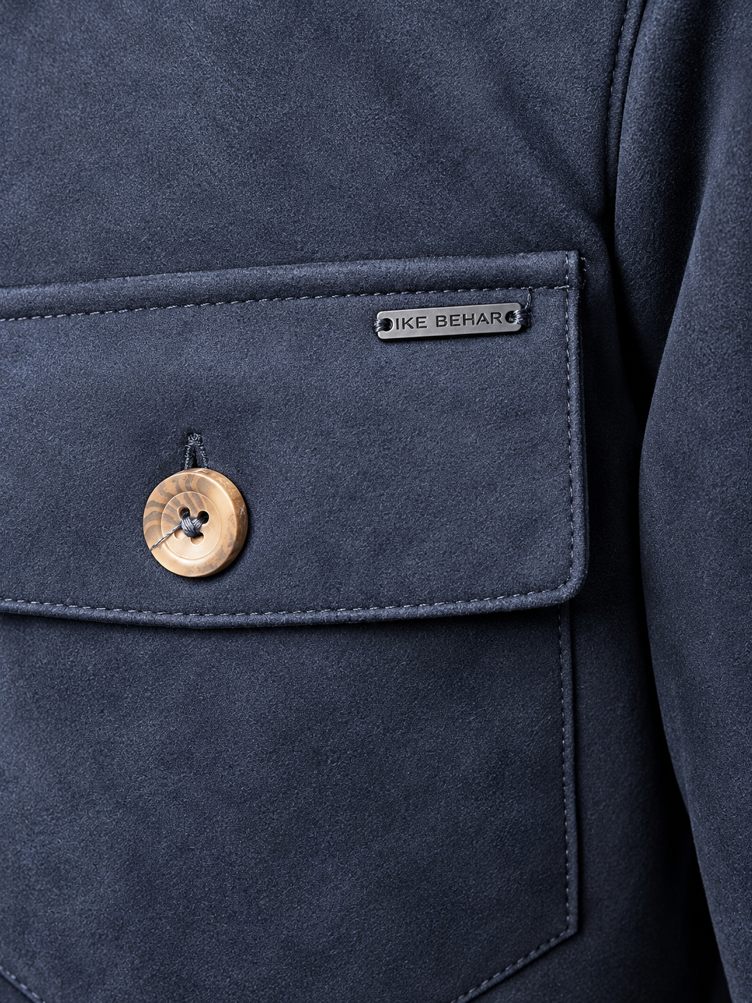 Close-up of a navy blue suede jacket pocket with a wooden button and a small metal brand plaque reading IKE BEHAR.