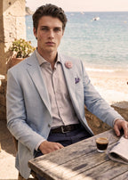 Man in light blue blazer sitting at a table by the sea with a newspaper and coffee.