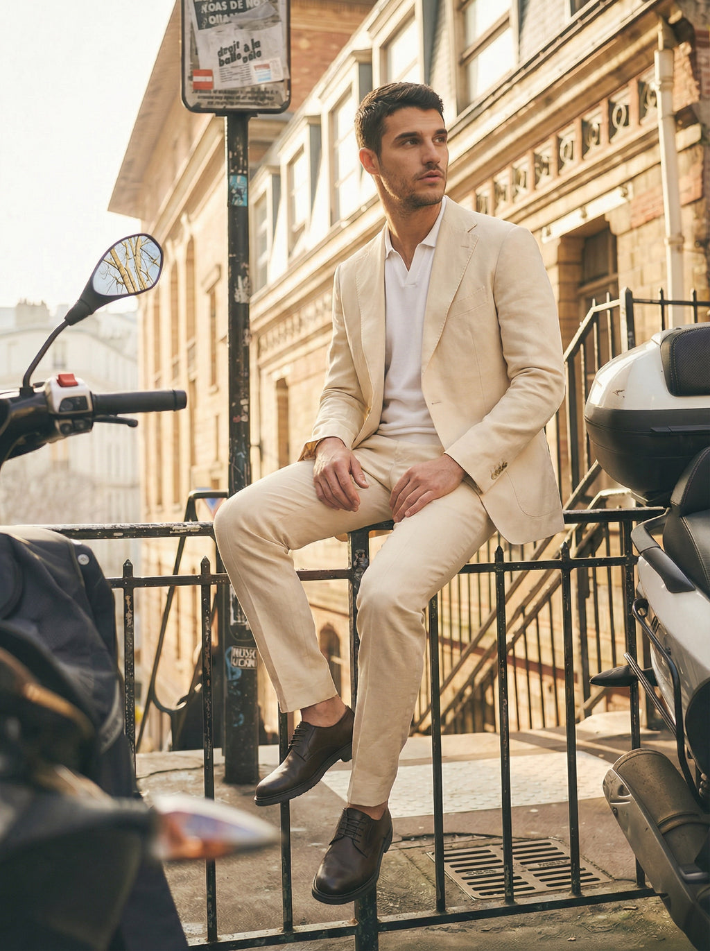 Man in a beige suit sitting on a railing with motorcycles in the foreground.