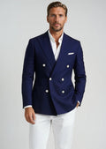 Man wearing a navy blue blazer with white buttons, white shirt, and white pants on a light gray background