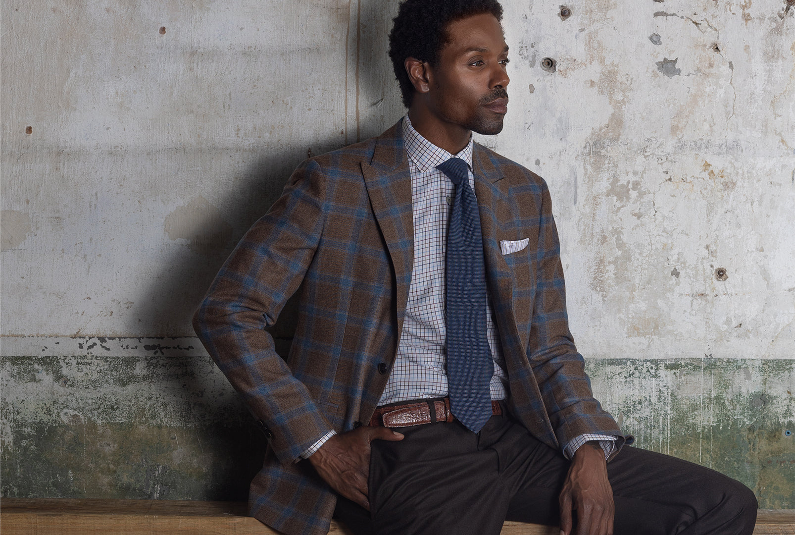 Model sitting on wooden bench against concrete wall wearing Ike Behar sport coat with hand in pocket