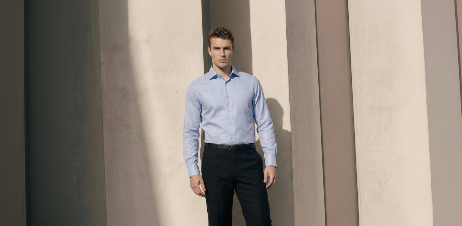 Man wearing a light blue dress shirt and black pants standing against a neutral wall.
