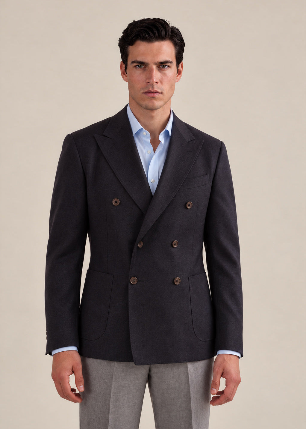 A man wearing a dark navy double-breasted blazer over a light blue dress shirt and gray trousers, standing against a solid beige background.
