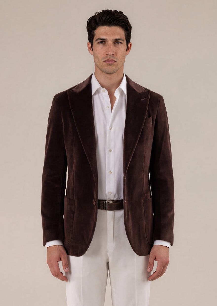 A man stands facing forward wearing a dark brown velvet blazer over a white button-down shirt, paired with white trousers and black leather loafers, against a plain beige studio background.