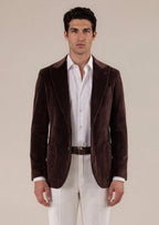 A man stands facing forward wearing a dark brown velvet blazer over a white button-down shirt, paired with white trousers and black leather loafers, against a plain beige studio background.