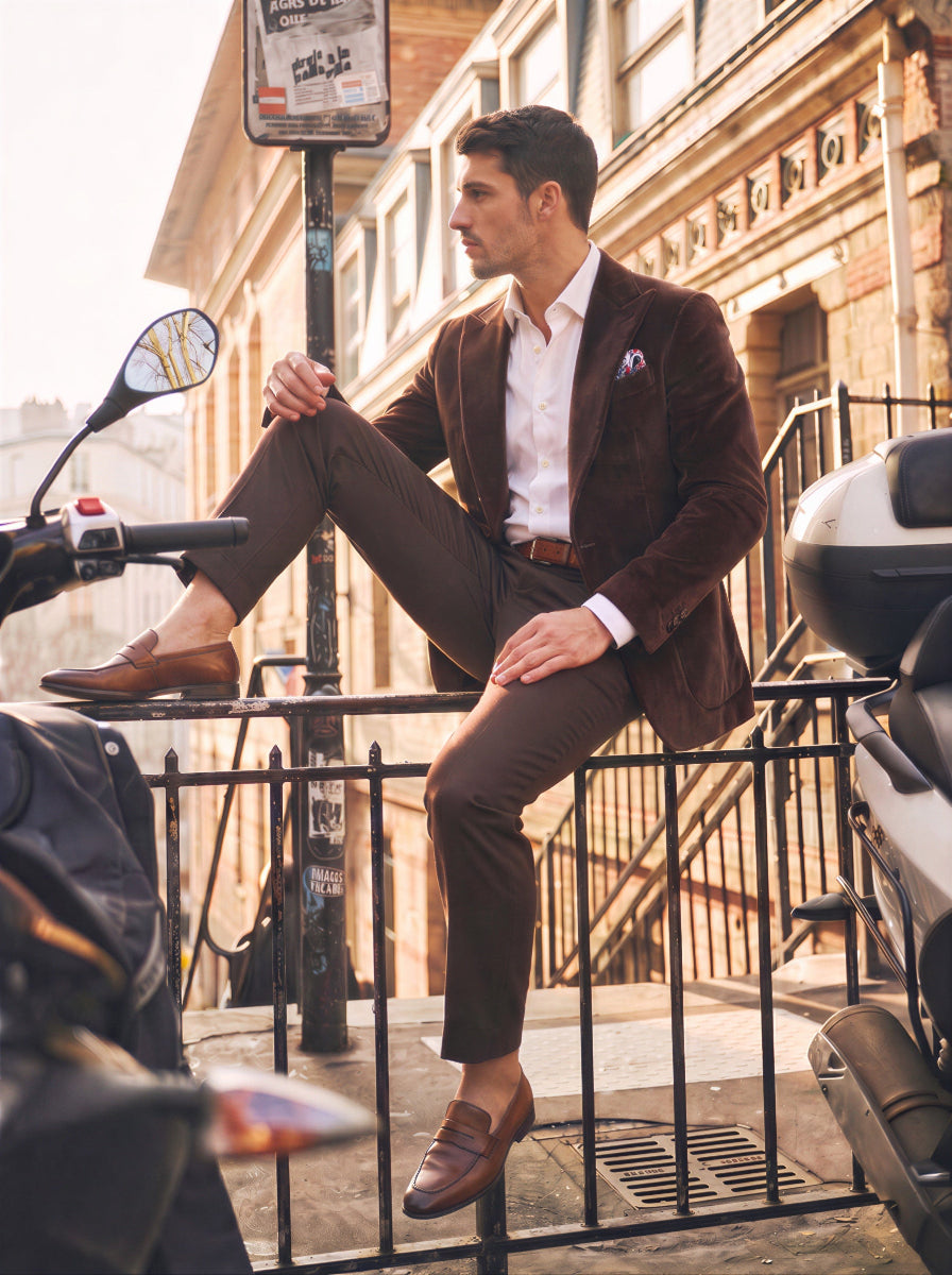 A man wearing a brown velvet suit, white dress shirt, and brown leather loafers sits casually on a black metal railing on a city sidewalk, with buildings and parked scooters in the background.