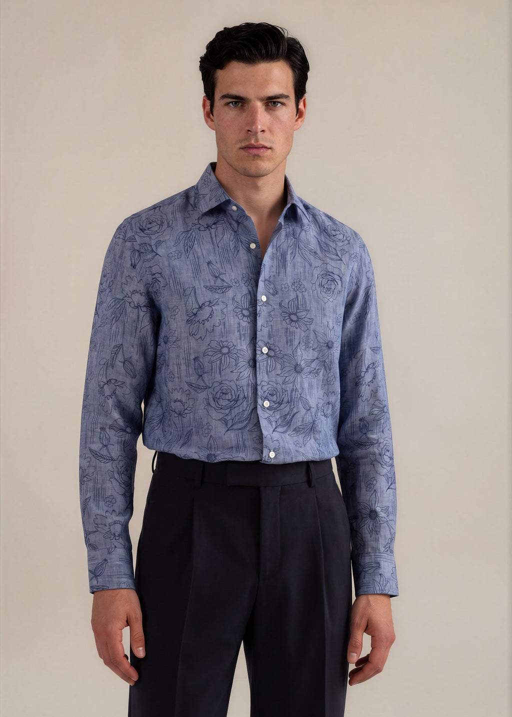 A man wearing a long-sleeve button-up shirt with a blue floral pattern, paired with dark trousers, standing against a plain beige background.