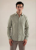 A man wearing a long-sleeve button-up shirt with a green and white gingham check pattern, paired with gray trousers, standing against a plain beige studio background.
