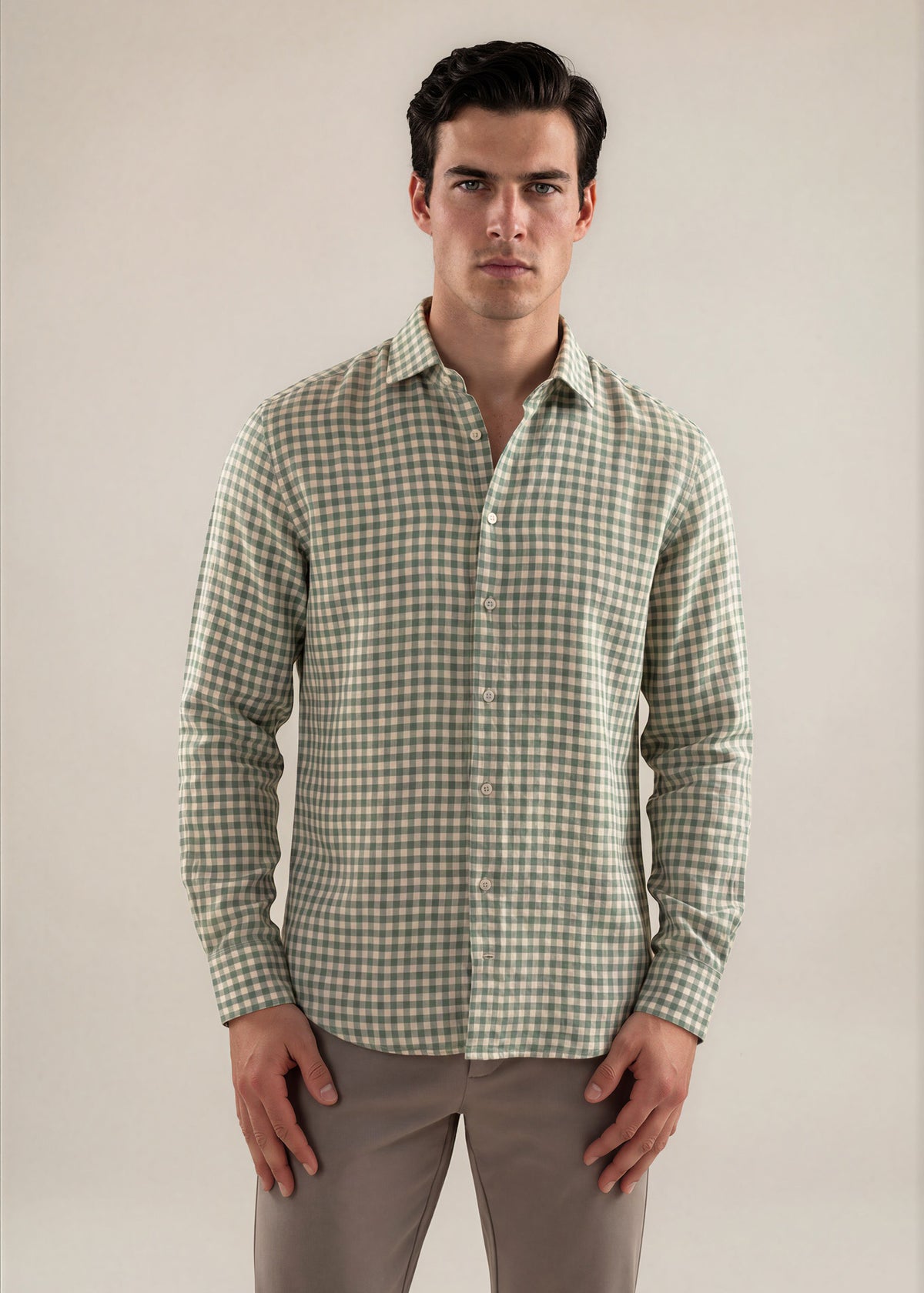 A man wearing a long-sleeve button-up shirt with a green and white gingham check pattern, paired with gray trousers, standing against a plain beige studio background.