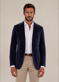 A man wearing a navy blue velvet blazer over a white button-down shirt and beige trousers, standing against a plain beige studio background.