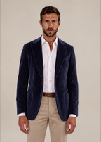 A man wearing a navy blue velvet blazer over a white button-down shirt and beige trousers, standing against a plain beige studio background.