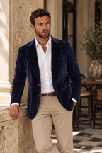 A man wearing a navy blue velvet blazer, white dress shirt, and beige trousers, leaning against a stone pillar in an outdoor patio setting.