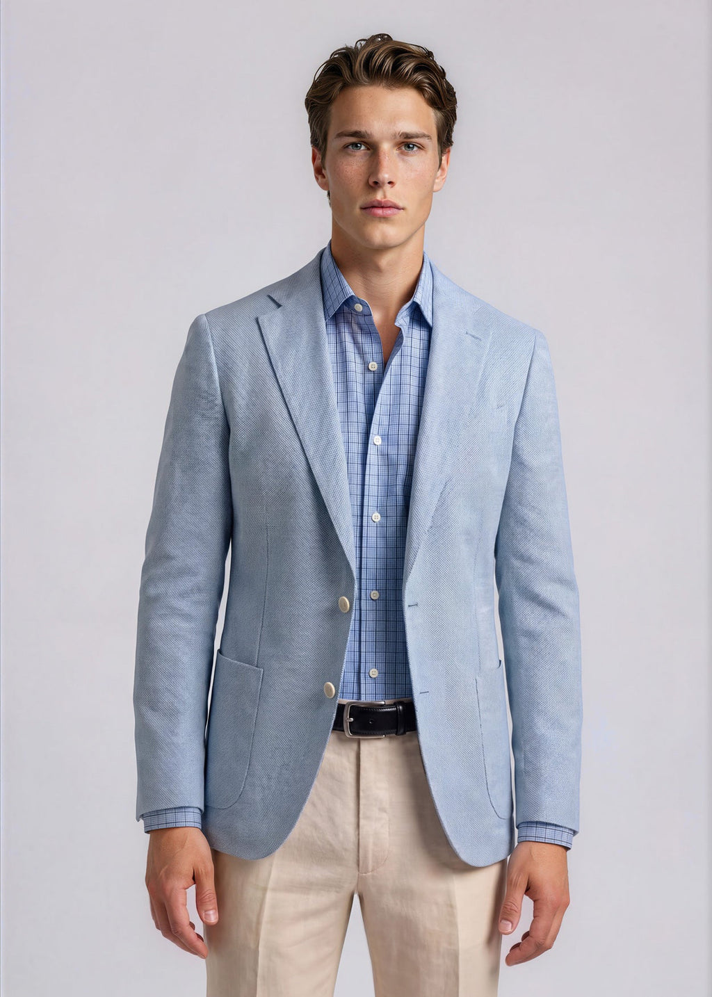 Man wearing a light blue blazer over a checkered shirt with beige pants on a plain background