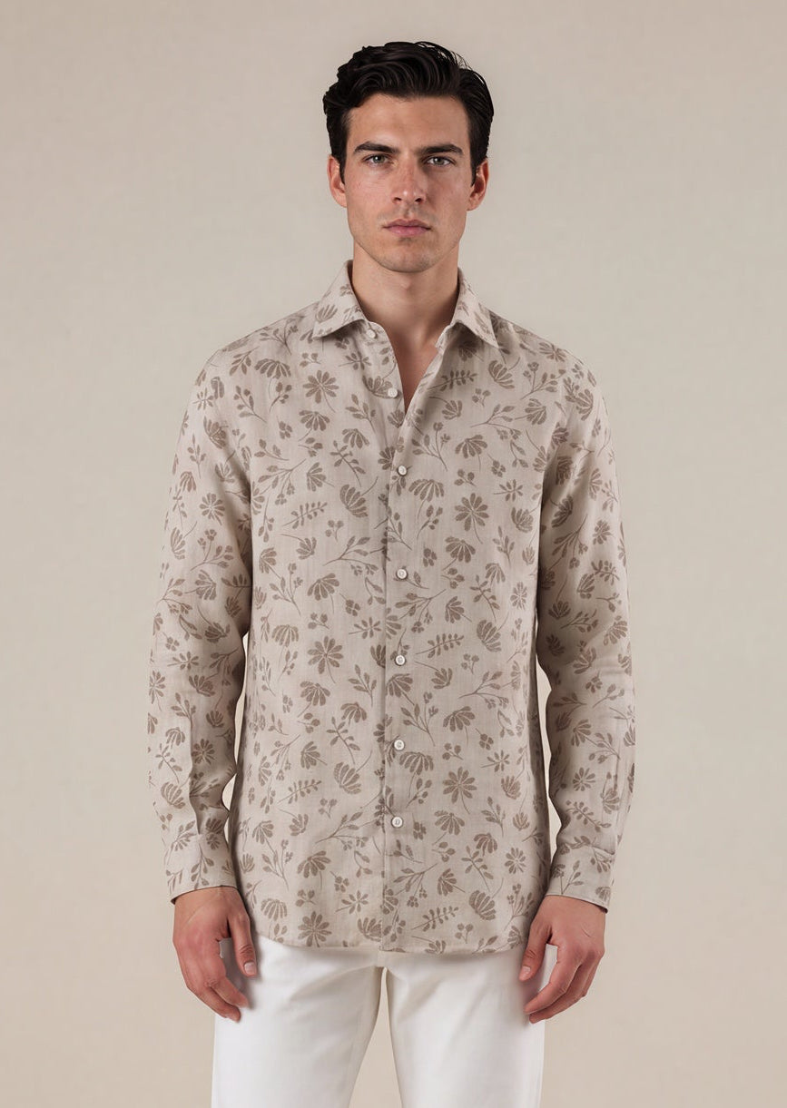 A man modeling a light beige long-sleeve button-up shirt with a brown floral pattern, paired with white trousers and olive green shoes, against a solid beige background.