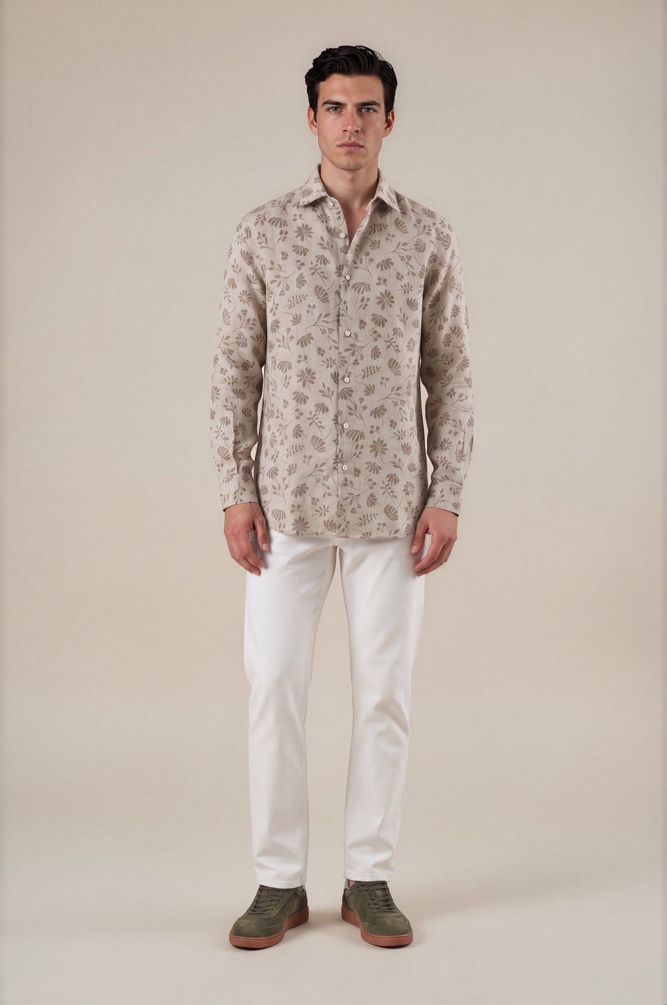 A man modeling a light beige long-sleeve button-up shirt with a brown floral pattern, paired with white trousers and olive green shoes, against a solid beige background.