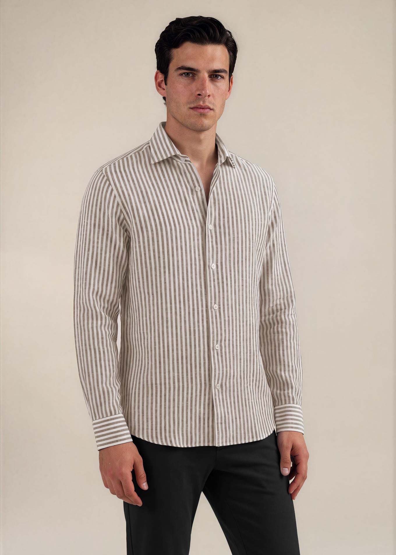 A man wearing a beige and white vertically striped long-sleeve button-down shirt with black trousers, standing against a plain beige studio background.