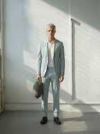 Man in light blue suit holding a bag against a white wall with sunlight filtering through.